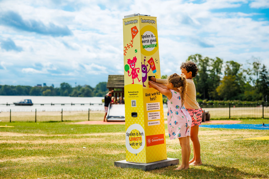 Zwei Kinder holen sich Sonnencreme an einer Sonnencreme-Station im Intertoys-Design auf einem Spielplatz am See