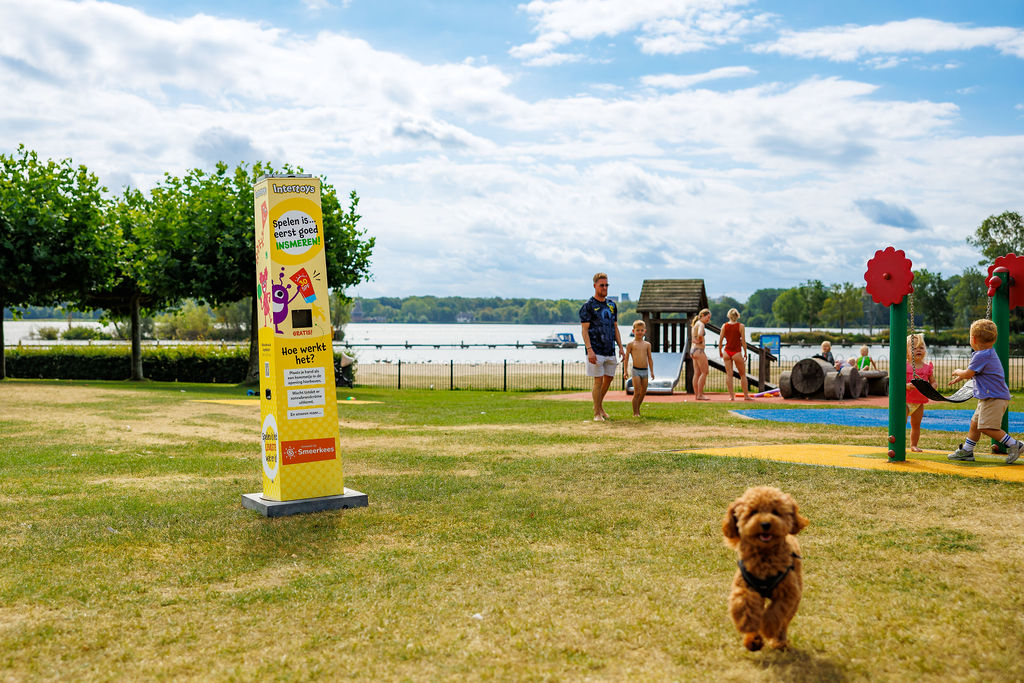 Gelbe Sonnencreme-Station im Intertoys-Design mit kostenloser Sonnencreme auf einem Spielplatz am Wasser, im Hintergrund spielende Kinder und ein Hund im Vordergrund