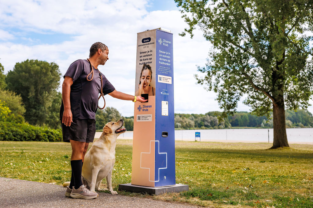 Mann mit Hund an einer Sonnencreme-Station der niederländischen Krankenkasse Zilveren Kruis in einem Park am See