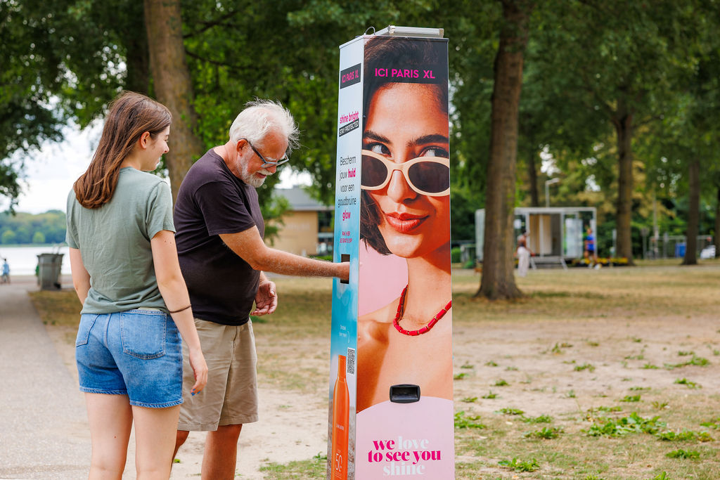 Älterer Mann hält seine Hand in die Öffnung einer Sonnencreme-Spendersäule von ICI PARIS XL mit Werbemotiv, daneben eine junge Frau, in einem Park mit Bäumen am Wasser