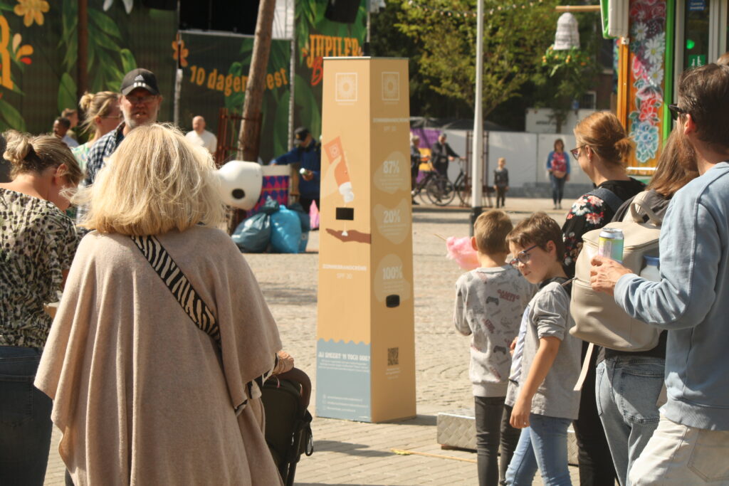 Beige Sonnencreme-Spendersäule von Smeerkees mit kostenloser Sonnencreme LSF 30 auf einem belebten Platz bei einem Stadtfest, umgeben von Besuchern, Familien und Kindern