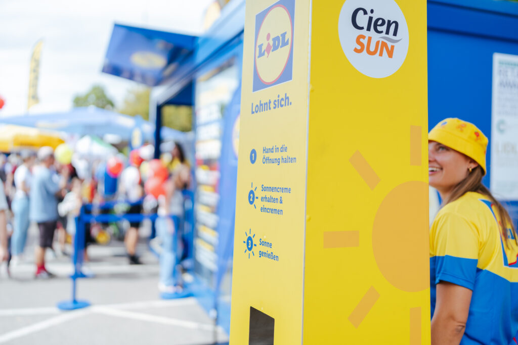 Nahaufnahme einer gelben Sonnencreme-Spendersäule von Lidl und Cien Sun mit der Anleitung „Hand in die Öffnung halten, Sonnencreme erhalten & eincremen, Sonne genießen", daneben eine lächelnde Promoterin mit gelbem Fischerhut, im Hintergrund ein Lidl-Stand mit Besuchern bei einer Veranstaltung im Freien