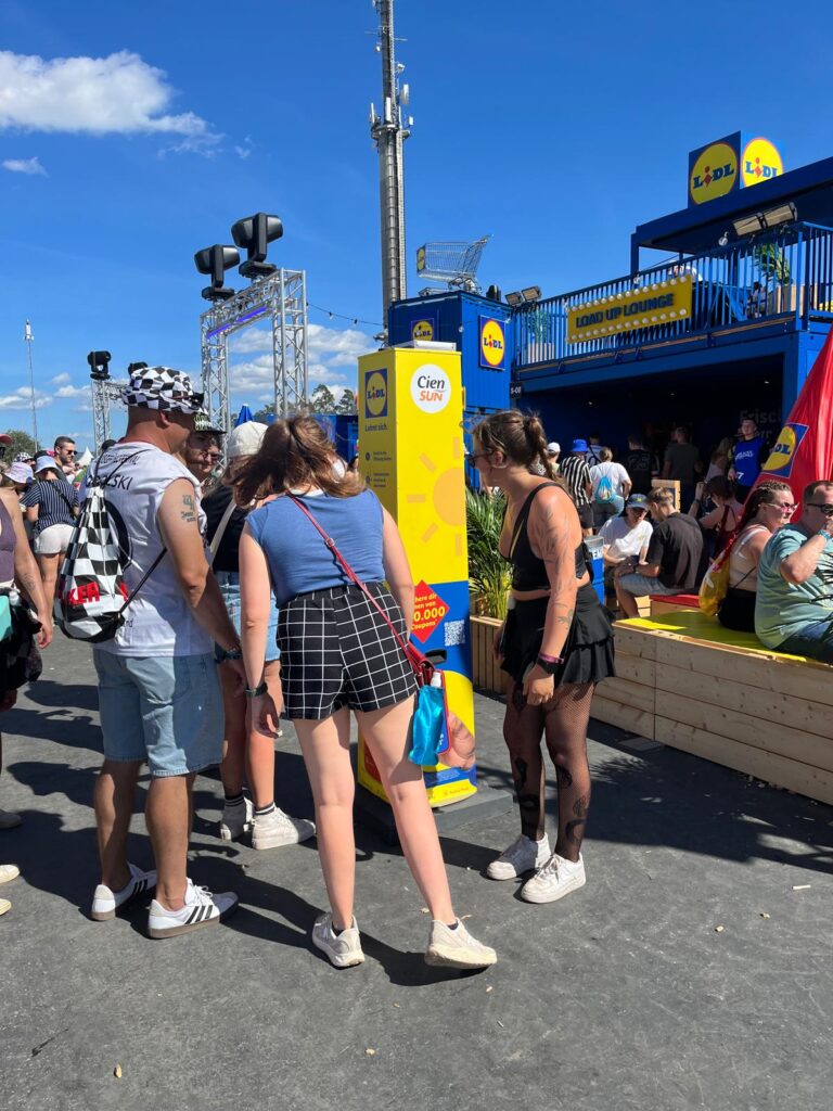 Junge Festivalbesucher stehen an einer gelben Sonnencreme-Spendersäule von Lidl und Cien Sun vor der Lidl „Load Up Lounge" auf einem sonnigen Festivalgelände